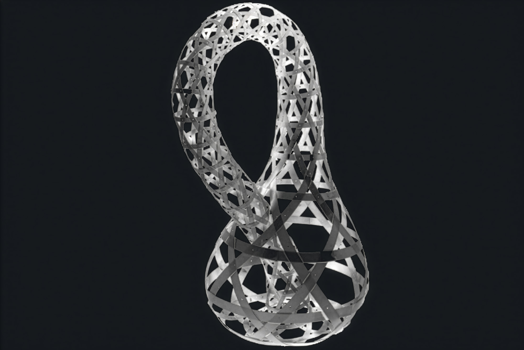 A woven looped surface of interlaced curved ribbons, a Klein-bottle-like form, against a dark background